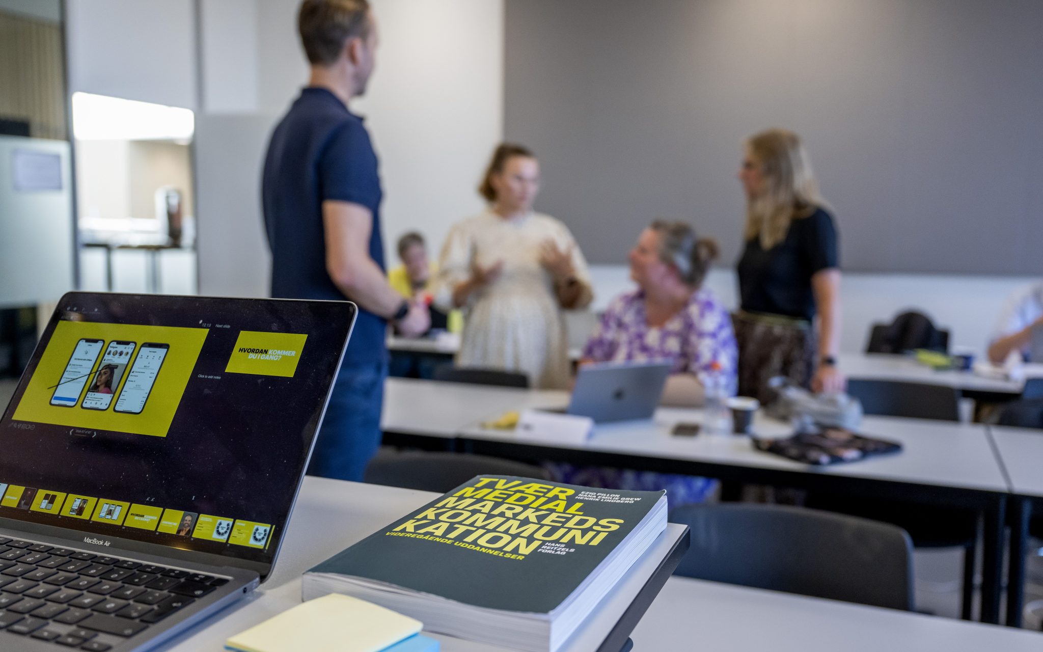 I forgrunden og i fokus er der en computer midt i et slideshow, og bogen "Tværmedial markedskommunikation". I baggrunden har vi et aktivt klasselokale med studerende.