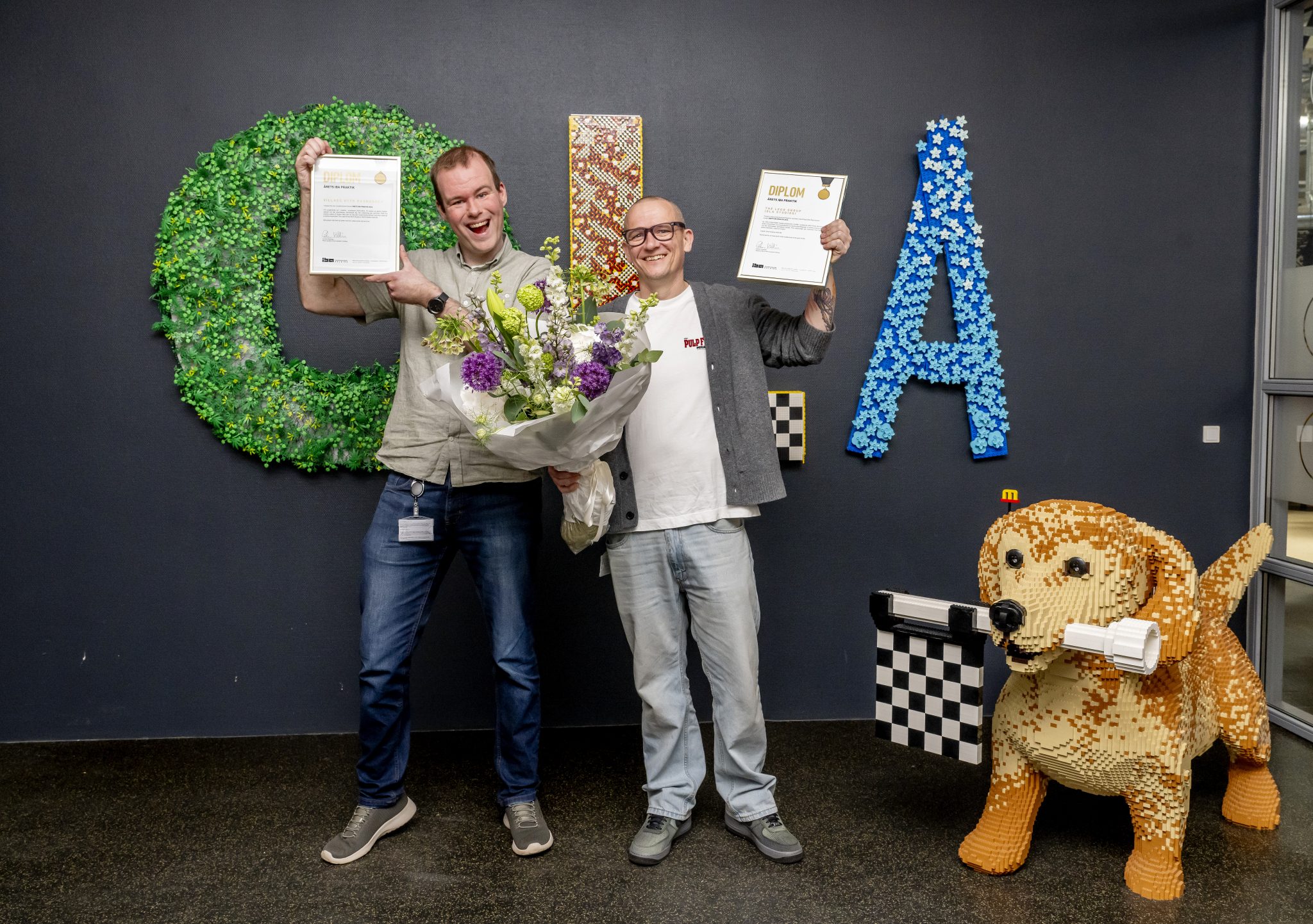 Foto af Villads og sin chef Søren, hos LEGO, der står og jubler med diplomer og blomster